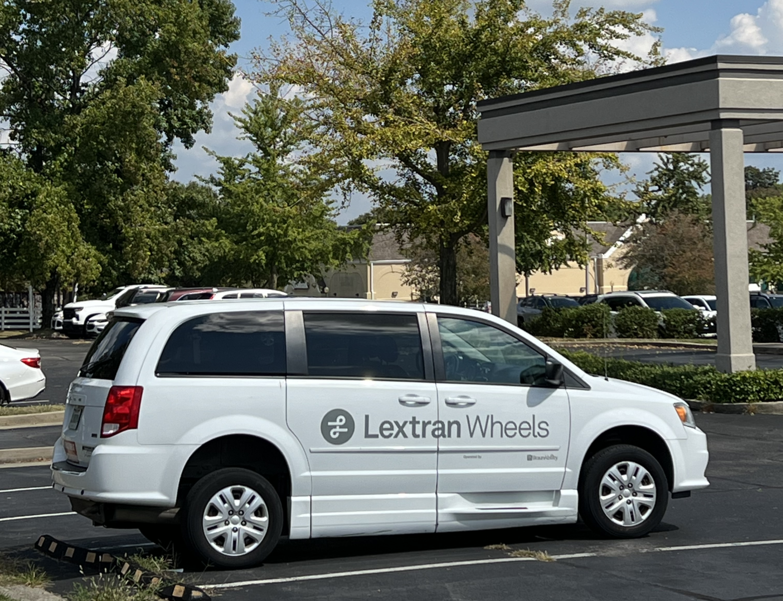 Image of a white paratransit van parked in a parking lot with the "Lextran Wheels" logo in gray along the side of the vehicle. The van is designed for accessible transportation, featuring additional space for passenger comfort and accommodations for individuals with disabilities.