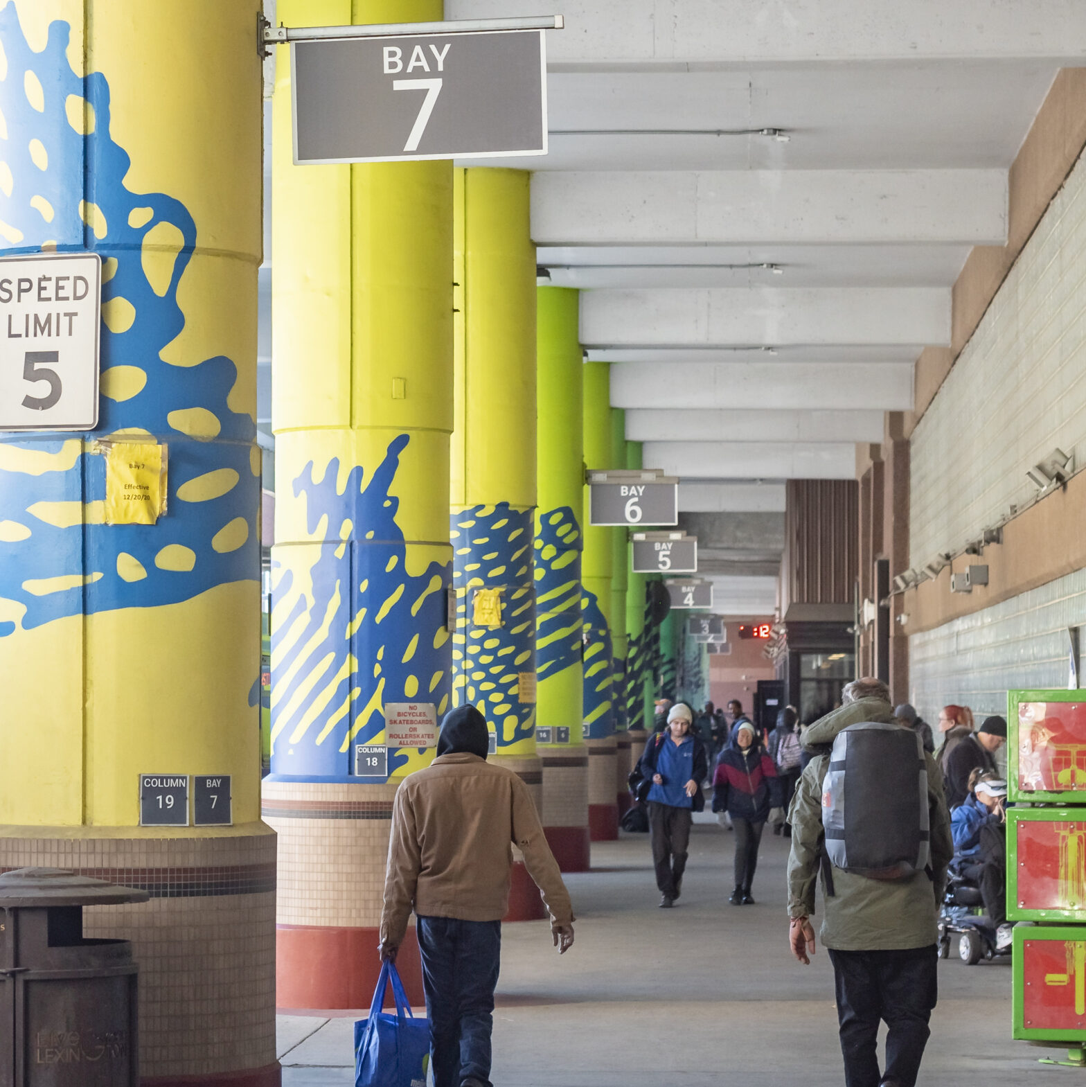 People walking through a brightly colored transit center with yellow and green pillars, marked with bay numbers, and signage indicating Bay 7. The area is covered, with various signs and a 5 mph speed limit sign visible. Passengers are carrying bags and wearing jackets, indicating a cold day.